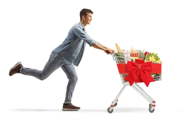 白い背景に隔離された赤いリボンと結ばれたショッピングカートで食べ物で走っている男性のティーンエイジャーの全長のプロフィールショット — ストック写真
