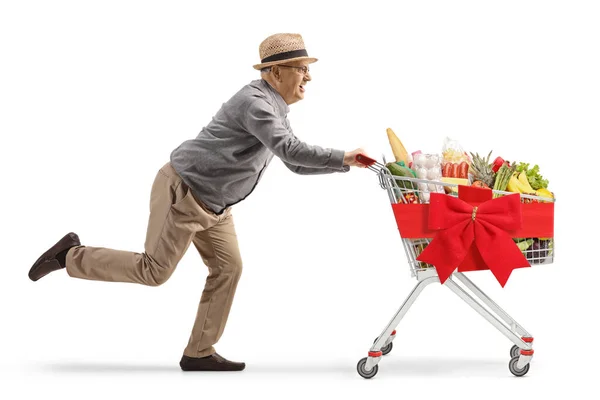 白い背景に隔離された赤い弓で飾られたショッピングカートで走っている高齢者の全長のプロフィールショット — ストック写真