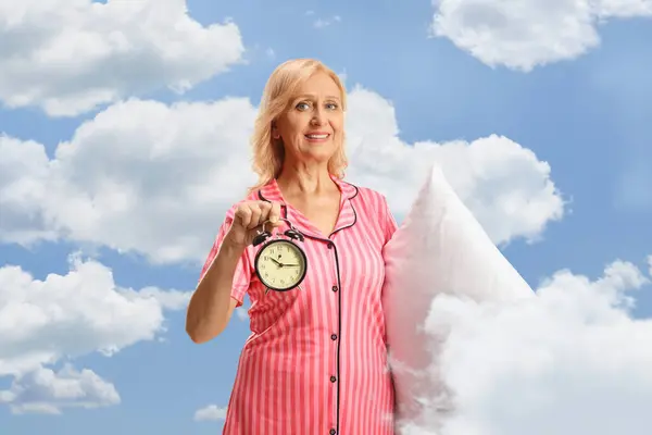 Mature woman in pajamas holding a pillow and alarm clock with clouds and sky in the background