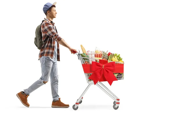 白い背景に隔離された赤いリボン弓と結ばれたショッピングカートで食べ物を持つ男性の学生の全長のプロフィールショット — ストック写真