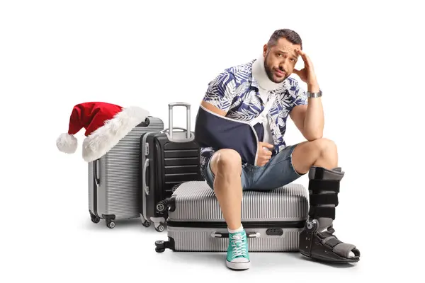負傷した腕と脚をスーツケースに座らせた悲しい男性の観光客 白い背景に隔離されたクリスマスの休日の概念 — ストック写真
