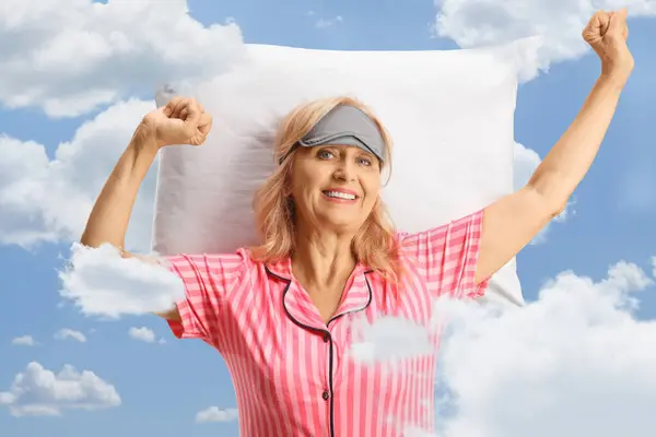 Happy mature woman in pajamas stretching on a pillow with clouds and sky in the background