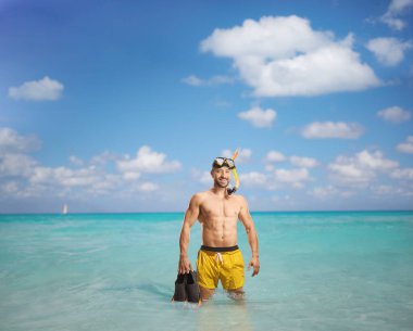 Dalış maskeli, yüzgeçlerini tutan ve denizde duran mayo giymiş bir adam.