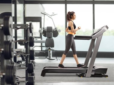 Spor salonunda koşu bandında yürüyen bir kadının tam boy profil fotoğrafı.