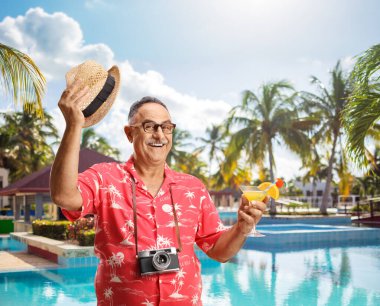 Olgun bir erkek turist elinde kokteyl ve yüzme havuzunun yanında şapka tutuyor.