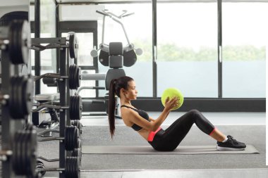 Paspasın üzerinde oturan ve spor salonunda topla egzersiz yapan genç bir kadının tam boy profil çekimi.