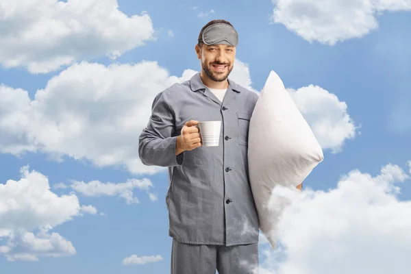 Man in pajamas and a sleeping mask holding a cup of coffee in front of clouds on a blue sky