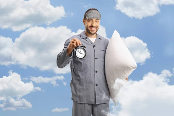 Man in pajamas with a sleeping mask holding a pillow and an alarm clock , clouds and blue sky in the background