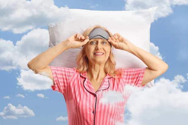 Woman in pajamas taking off sleeping mask with clouds and blue sky as a background