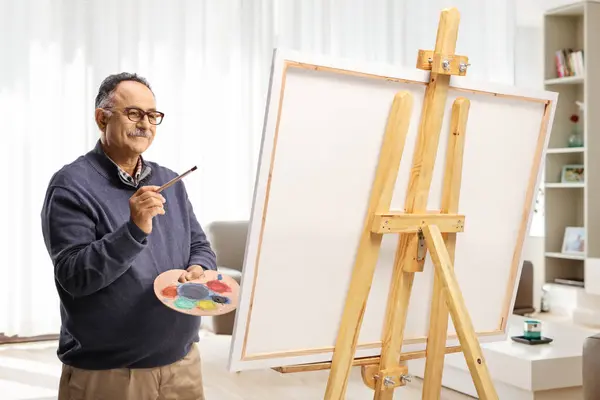 Mature man with a brush and palette painting on a canvas in an aparmtent