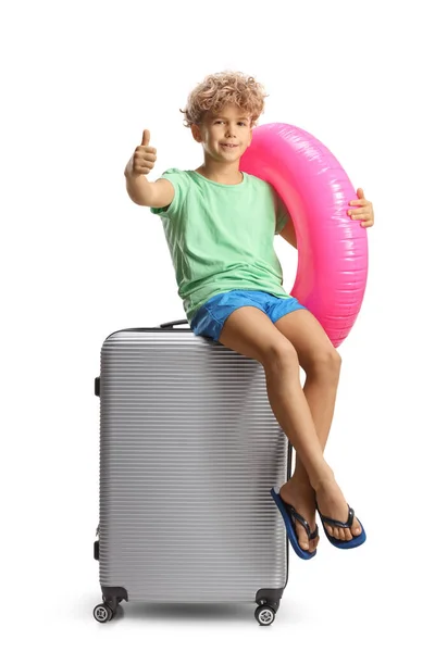 スーツケースに座って 白いアバックグラウンドで隔離された親指をジェスチャーするスイミングリングを持つ少年 — ストック写真