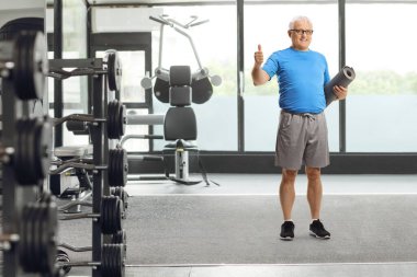 Spor kıyafetli, spor kıyafetli, paspas tutan ve spor salonunda baş parmağını gösteren olgun bir adamın tam boy portresi.