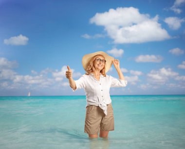 Hasır şapkalı orta yaşlı bir kadın denize daldırıyor ve baş parmağını kaldırıyor.