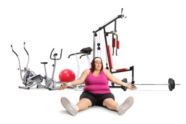 Tired plus size woman in sportswear sitting on the ground in front of a fitness gear solated on white background