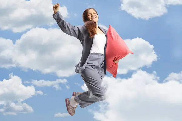 Young happy female in pajamas holding a pillow and jumping in the clouds