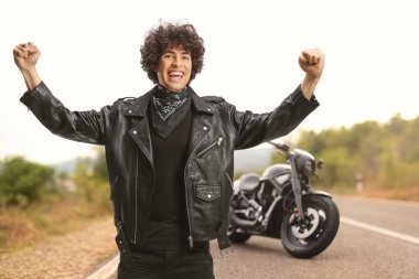 Deri ceketli ve siyah eşarplı heyecanlı genç motosikletçi yolda mutluluğu işaret ediyor. 