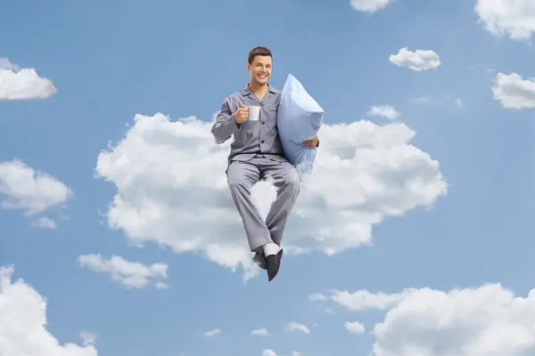 Young man in pajamas holding a cup and a pillow seated on a cloud up in the sky