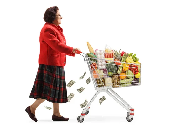 白い背景に隔離された食べ物とお金を持つショッピングカートで歩く高齢女性の全長のプロフィールショット — ストック写真