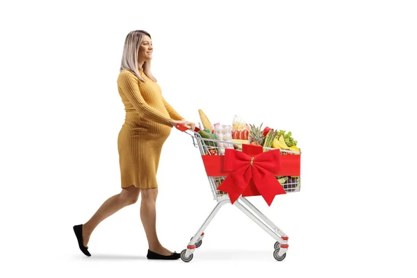 一名孕妇推着一辆装有健康食品的购物车 车上绑着一条白色背景的红丝带 照片全景拍摄 — 图库照片