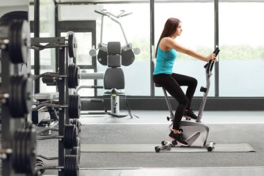 Spor salonunda bisiklete binen genç bir kadının tam boy profil fotoğrafı.