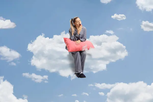 Full length shot of a young sleepy woman sitting on a cloud with a pillow and yawning