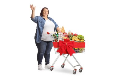 Dolu bir alışveriş arabasıyla duran ve beyaz arka planda izole bir şekilde el sallayan tombul bir kadının tam boy çekimi.