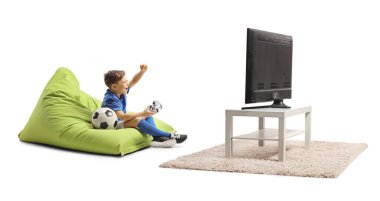 Çocuk televizyonun önünde armut çuvalında oturuyor ve beyaz arka planda izole edilmiş joystick ile futbol oynuyor.