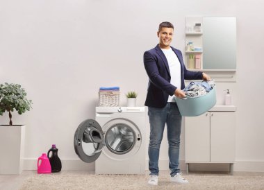 Çamaşır sepetini banyoda kıyafetlerle tutan genç bir adamın tam boy portresi.