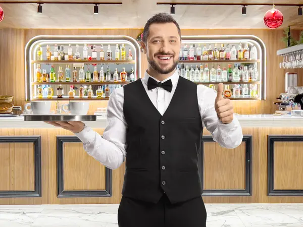 Full length portrait of a waiter holding a tray with coffee cups and showing thumbs up at a bar
