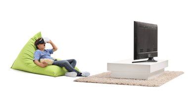 VR gözlüklü bir çocuk patlamış mısır yiyor ve beyaz arka planda izole edilmiş bir televizyonun önünde puf minderin üzerinde oturuyor.