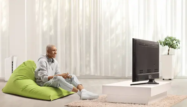 ハッピーアフリカ系アメリカ人が緑色のビーンバッグに座って 自宅でジョイスティックでビデオゲームをする — ストック写真