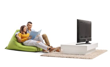 Genç bir çift, beyaz arka planda izole edilmiş bir televizyonun önünde oturup kitap okuyor.