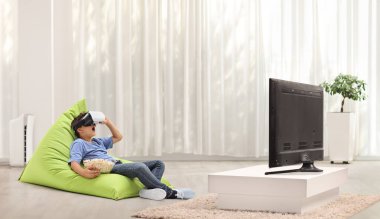 VR gözlük takan ve evde televizyonun önünde bir puf minder üzerinde oturmuş patlamış mısır yiyen bir çocuk.
