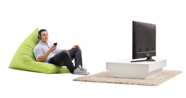 Armut koltukta oturan ve beyaz arka planda izole edilmiş televizyon karşısında kulaklıkla müzik dinleyen genç bir adam.