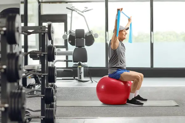Elderly man exercising with elastic band and a fitness ball at a gym