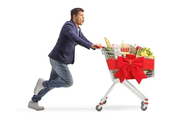 白い背景に隔離された赤いリボンと結ばれたスーパーマーケットのショッピングカートで走っている興奮した男のフル長さのプロフィールショット — ストック写真
