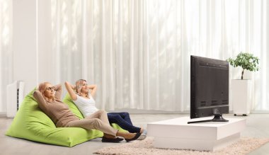 Evde iki kadın televizyon karşısında puf mindere oturuyor.