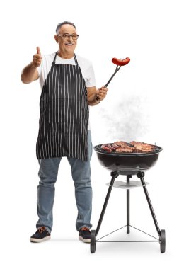 Olgun bir adam barbekü ızgarasında et pişiriyor ve beyaz arka planda izole edilmiş bir çatalla sosis tutuyor.