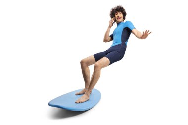 Dalgıç kıyafetli genç adam sörf tahtasına biniyor ve beyaz arka planda izole edilmiş akıllı telefon kullanıyor.