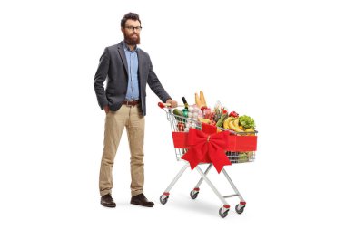 Beyaz arka planda kırmızı kurdeleli bir alışveriş arabasıyla poz veren sakallı bir adamın tam boy portresi.