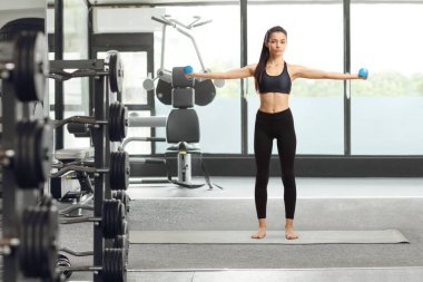 Siyah bluzlu genç bir kadının uzun bir portresi ve spor salonundaki bir paspasın üzerinde halterlerle egzersiz yapan taytlar.