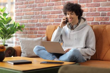Kafedeki bir kanepede dizüstü bilgisayarı ve akıllı telefonu olan genç bir adam.