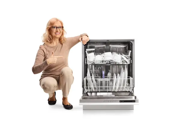 Middle Aged Woman Kneeling Next Open Dishwasher Pointing Clean Plates lizenzfreie Stockbilder