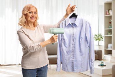 Askıda gömlek tutan ve evde buharlı ütü kullanan bir kadın.