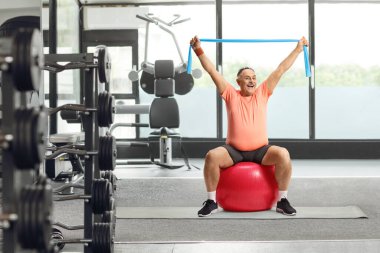 Spor giyim kuşağında gülümseyen olgun bir adam spor salonunda spor topunda oturup lastik bir bantla egzersiz yapıyor.