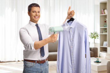 Evdeki oturma odasında gömleği için buharlı ütü kullanan mutlu genç adam.