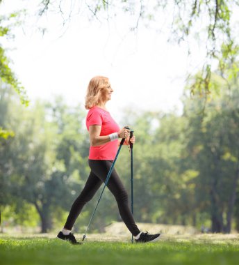 Orta yaşlı bir kadının parkta yürüyüş direğiyle yürüdüğünü gösteren uzun bir profil fotoğrafı.