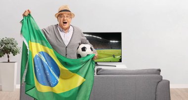 Heyecanlı bir futbol fanatiği evde bir kanepe ve televizyon önünde Brezilya bayrağı tutuyor.