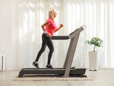Evinde koşu bandında koşan orta yaşlı bir kadının tam boy profil fotoğrafı.