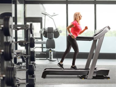 Orta yaşlı bir kadının spor salonundaki koşu bandında koşarken tam boy profil fotoğrafı.